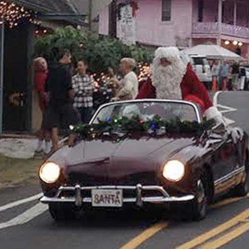 A classic maroon convertible driving Santa Claus down a street lined with holiday lights and onlookers, as a festive parade scene unfolds.