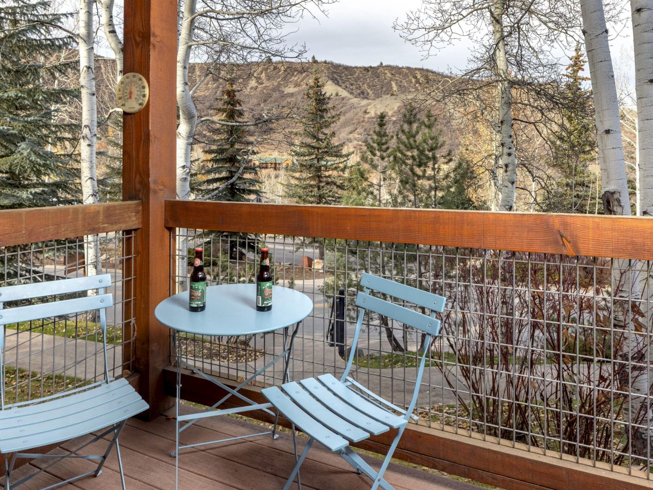 A small balcony with light blue metal table and two chairs, three bottles on the table, wooden railing, trees and hills in the background, peaceful outdoor scene.