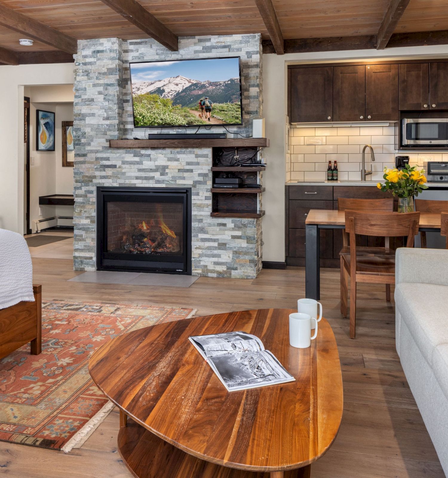 Cozy living area with a stone fireplace, mounted TV, open kitchen, wooden beams, and a round coffee table in front of a sofa. End the sentence with a period.