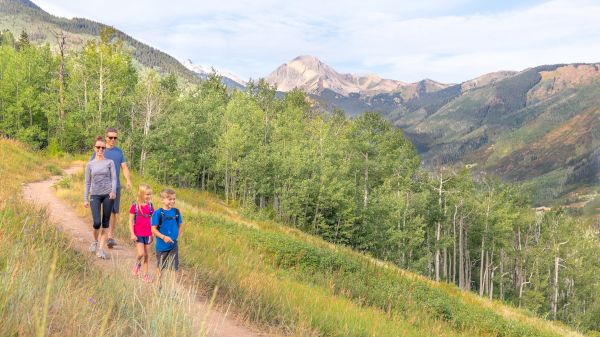 A family hike on a mountain trail with a man and two children walking along a sunny hillside forest path, mountains in the background.