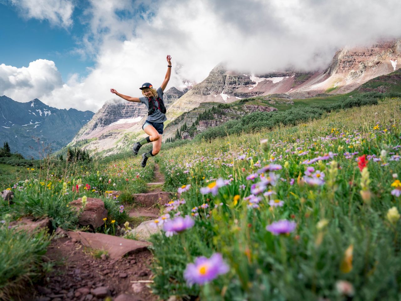 A person leaps joyfully on a rocky trail in a wildflower-filled mountain meadow, with rugged peaks and blue sky in the background.