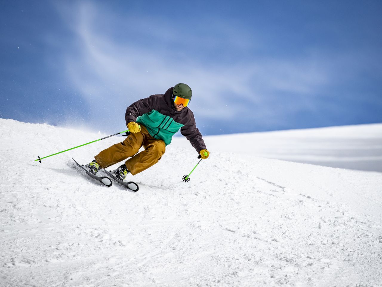 A skier wearing a green and black jacket, brown pants, and a green helmet carving down a snowy slope with bright blue sky behind.