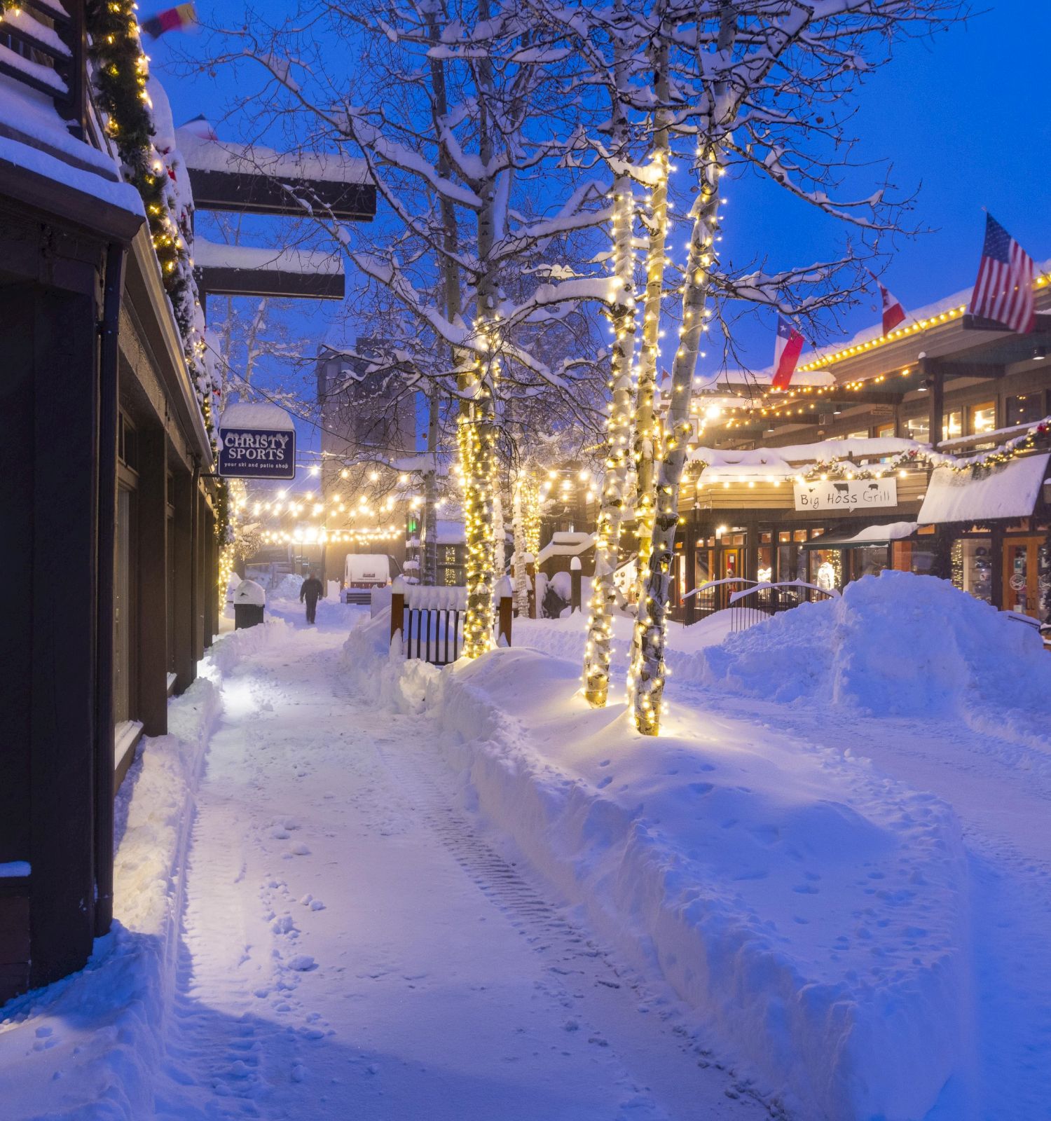 A snowy village street at dusk with twinkling lights on trees, shop windows, and shoveled paths lining a cozy, festive town square.
