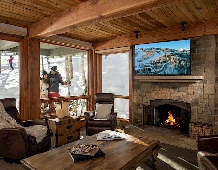 A cozy living room with a stone fireplace, a mounted TV, wooden beams, and a couch set; people outside socializing near large windows.