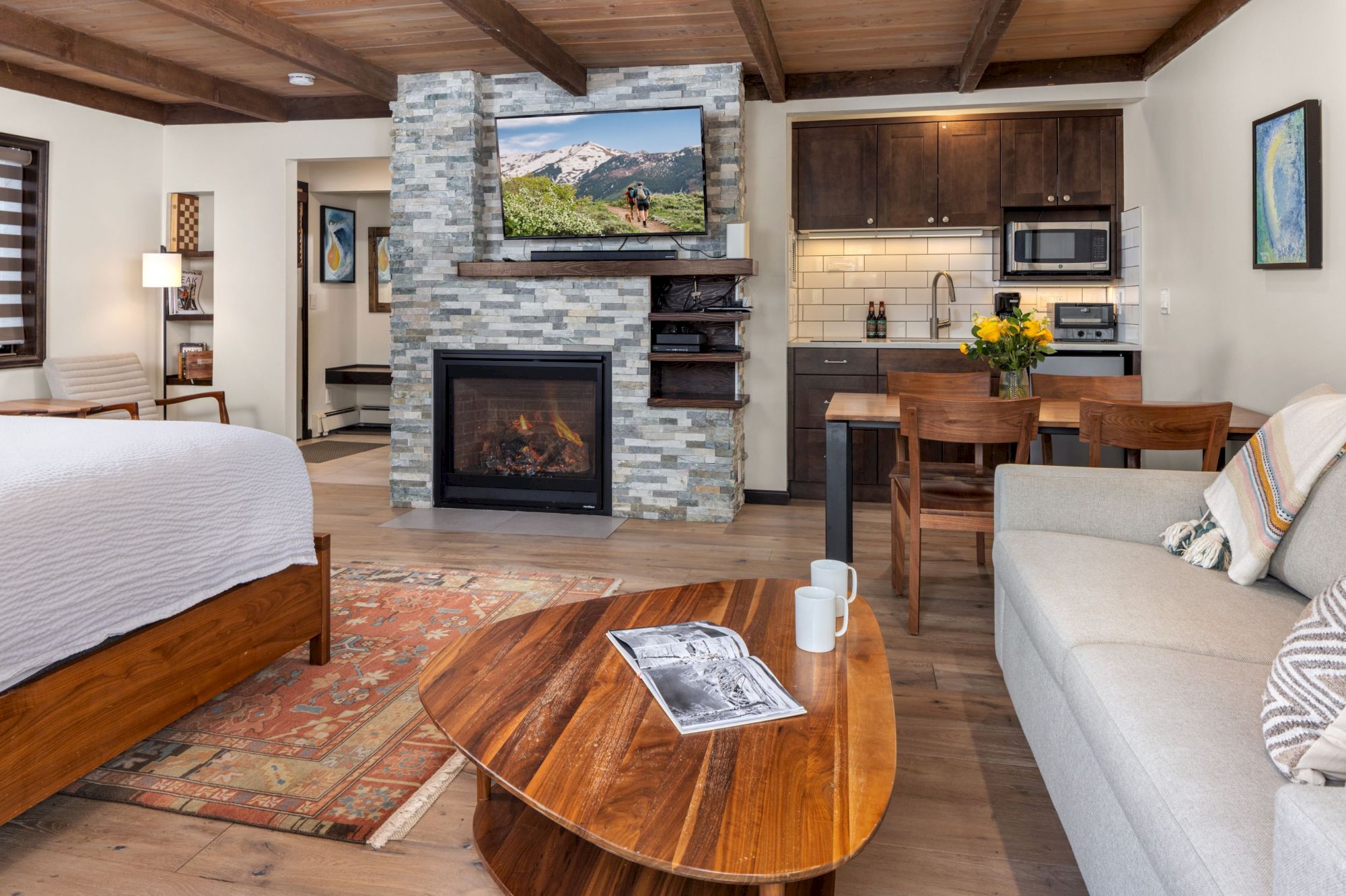 A cozy living area with a stone fireplace, TV above, kitchenette, and a wooden coffee table; a sofa and bed, warm lighting, and a rug.