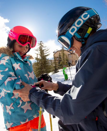 Two skiers in a snowy resort; one helps adjust the other&rsquo;s gear while wearing helmets and goggles, blue sky and trees in background.