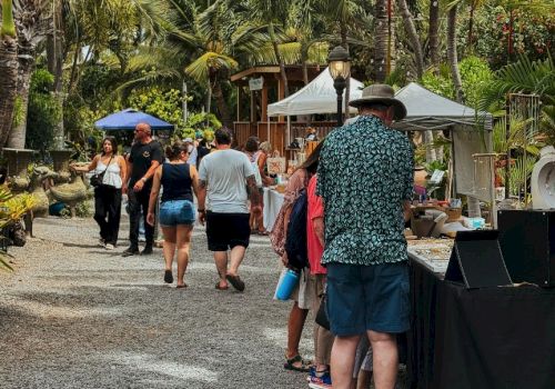 A sunny outdoor market with people browsing stalls, tropical trees behind, and casual shoppers in summer clothes at booths by the path.