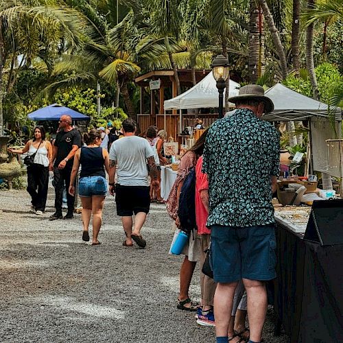 A sunny outdoor market with people browsing stalls, tropical trees behind, and casual shoppers in summer clothes at booths by the path.