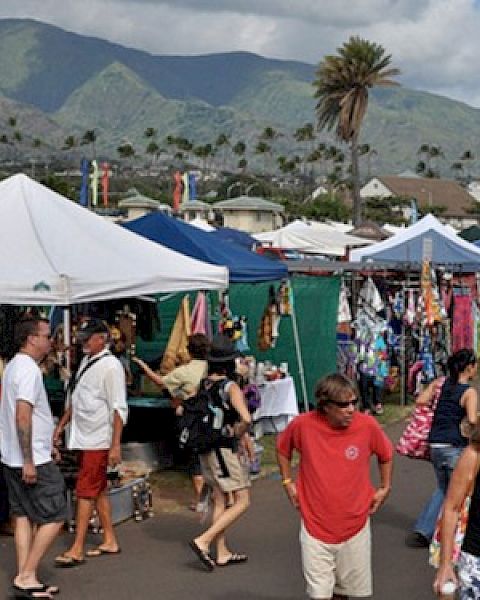 A bustling outdoor market with white tents, vendors selling goods, shoppers strolling, and a mountain backdrop under a sunny sky. (ends with period)