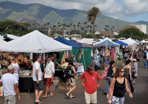 A bustling outdoor market with white tents, vendors selling goods, shoppers strolling, and a mountain backdrop under a sunny sky. (ends with period)