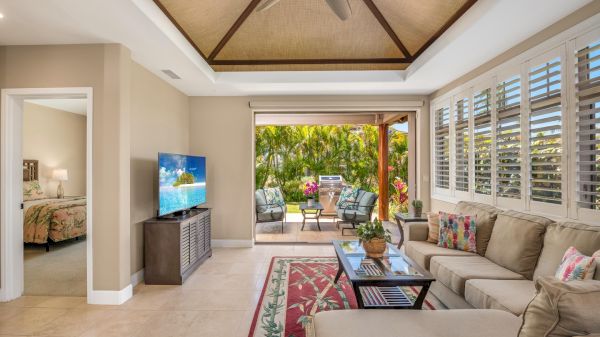 A bright living room with beige sofa, glass coffee table, TV on a stand, and open sliding doors to a sunny outdoor patio; tropical plants outside.