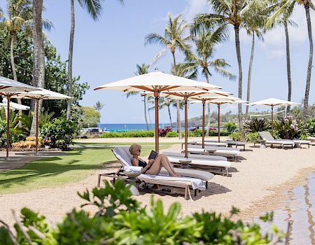 A tropical beach scene with palm trees, lounge chairs under umbrellas, a person relaxing on a sunbed, and calm water in the background.