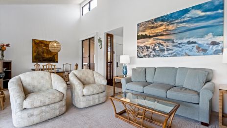 A cozy, airy living room with a blue sofa, two beige armchairs, and a glass coffee table, plus ocean-themed wall art.