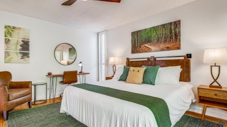 A cozy hotel room with a large bed, green accents, wooden furniture, a ceiling fan, and a seating area with a chair and desk by the window.