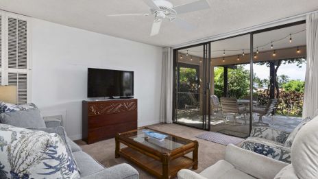 A bright living room with a gray sofa, coffee table, TV on a wooden stand, ceiling fan, and sliding glass doors to a patio.