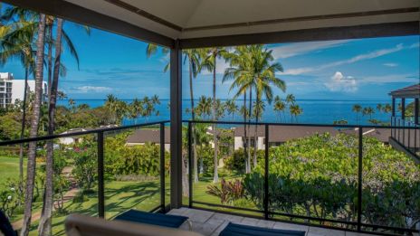 A balcony overlooks a tropical resort with palm trees, lush greenery, and a clear blue ocean beyond, under a bright sky.