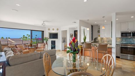 A bright, open-concept living space with a kitchen, dining area, and living room, large windows, bar seating, and tropical outdoor view.