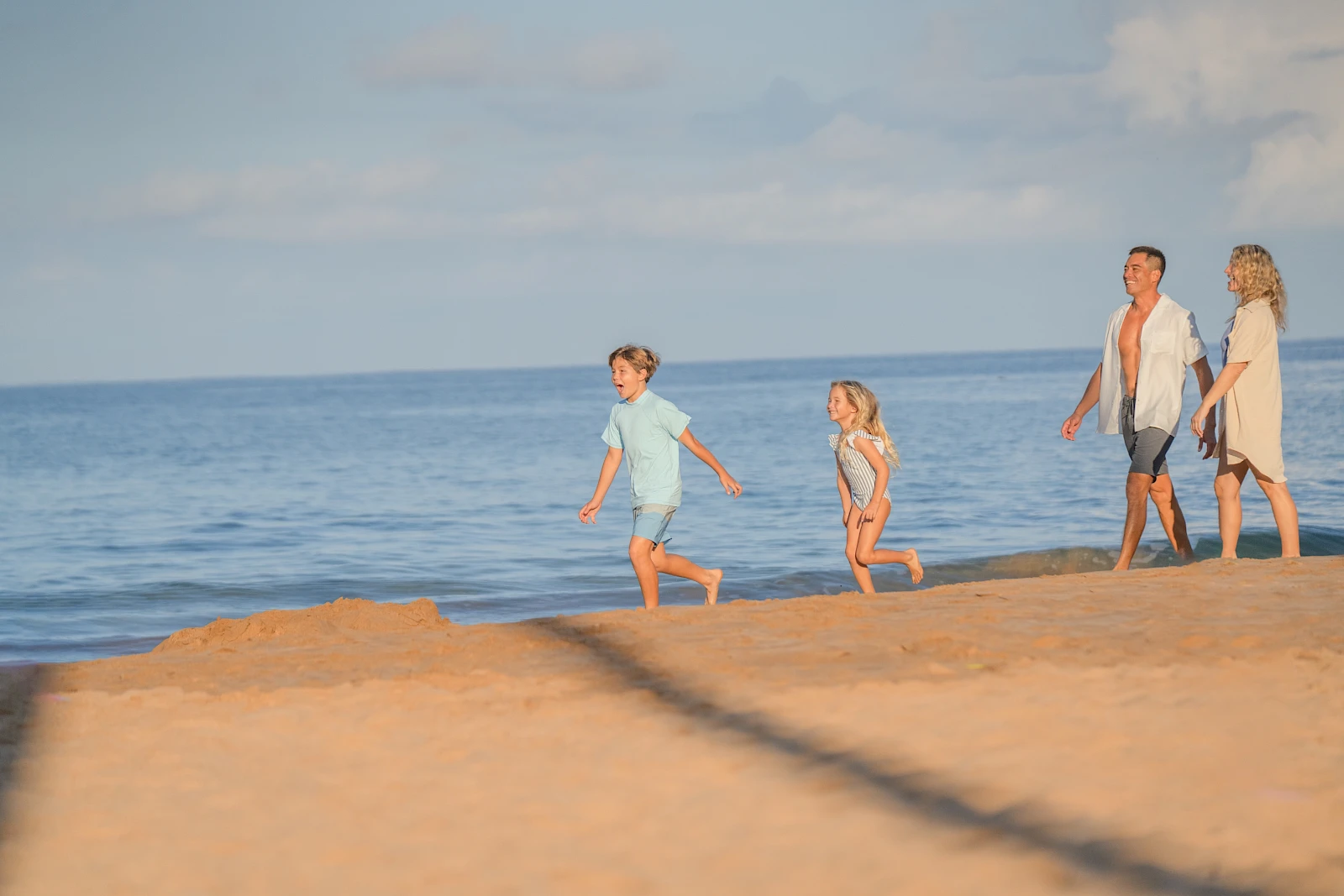 A family of four walks along a sandy beach by the calm sea, children leading toward the water as the parents follow, sunny day.