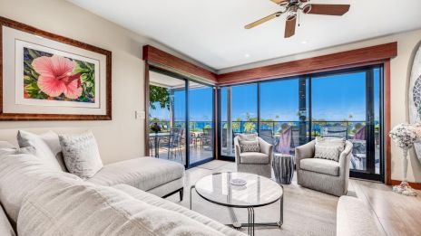 A bright living room with light sofa, glass coffee table, wicker chairs, large sliding glass doors to a balcony, ocean view, ceiling fan, tropical decor.