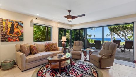 A bright living room with a beige sofa and two armchairs, a round rug, glass coffee table, tropical art, and a sliding door to a sunny balcony with ocean view.