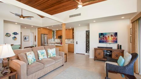 A bright open living area with a tan sofa, tropical throw pillows, wicker chair, wooden ceiling, and a view into the kitchen; wall art and a TV console complete the space.