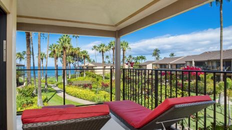 A balcony with red cushioned lounge chairs overlooks a tropical garden and palm trees, with a lake and blue sky beyond the railing.