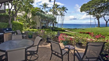An outdoor patio with metal chairs and tables, tropical landscaping, palm trees, and a view of a blue ocean under a partly cloudy sky.