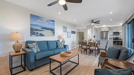 A bright, open living-dining area with blue couches, wooden coffee and side tables, coastal artwork, ceiling fans, and a modern kitchen in the background.