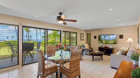 Bright open living area with ceiling fan, tiled floor, and large sliding doors to a balcony; dining table, wicker chairs, and a cozy seating nook, overlooking greenery and water.