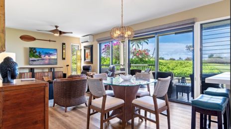 A bright, open dining-living area with a round dining table and six chairs, large sliding glass doors to a patio, tropical plants outside, ceiling fan, and a cozy seating cluster near a wall-mounted TV.