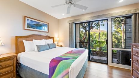 A bright hotel or bedroom with a large bed, wooden nightstands, ceiling fan, and glass doors opening to a balcony with greenery outside.