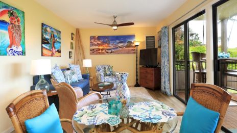 A bright tropical living room with blue and teal accents, colorful art on yellow walls, wicker furniture, and a sliding door to an outdoor deck.