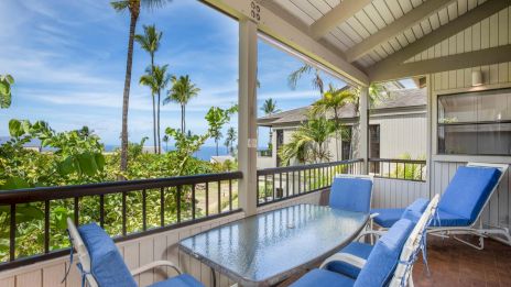 A sunny balcony with blue cushioned chairs, a glass table, palm trees, and a ocean view in the distance.