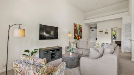 Cozy living room with light gray sectional, colorful patterned chairs, wall-mounted TV, modern fireplace, round coffee table, plants, and soft ambient lighting.