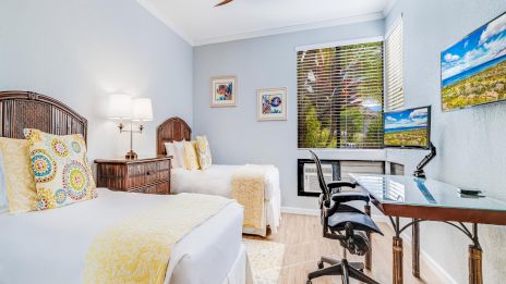 Two twin beds in a bright blue hotel room with a desk, chair, wall-mounted TV, window blinds, and a ceiling fan.