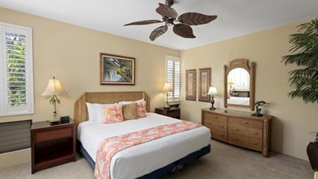 A cozy bedroom with a king bed, wooden nightstands, matching dresser, tropical ceiling fan, warm beige walls, and plants by a large dresser.