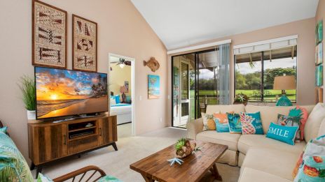 A bright, tropical-style living room with a beige sofa, teal accents, a wooden coffee table, a TV stand, potted plants, and a sliding door opening to a sunny outdoor view.