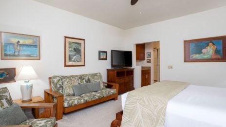 A bright hotel room with a bed, a floral sofa set, a dresser, TV, nightstand lamps, and framed beach-themed paintings on white walls.