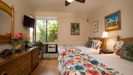 A cozy hotel room with two double beds in floral bedding, wood dresser, wall art, a TV, lamps, plants, and a windowed view&mdash;bright and inviting.