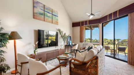 A bright living room with beige furniture, a wall-mounted TV, and a glass coffee table. Sliding doors lead to a sunny balcony with ocean views.