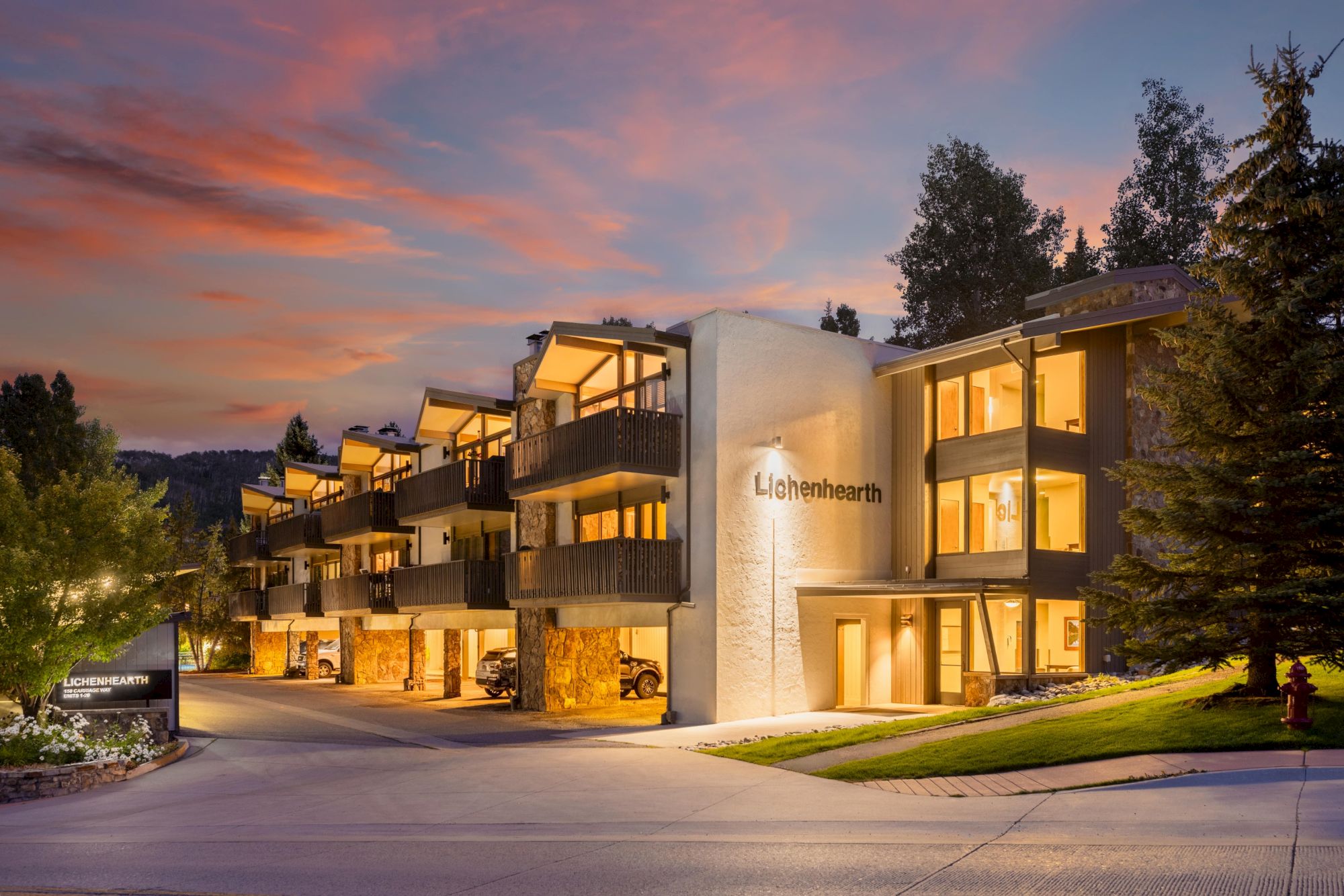 The image shows a modern hotel building at dusk, featuring balconies, landscaped surroundings, and a colorful sunset sky.