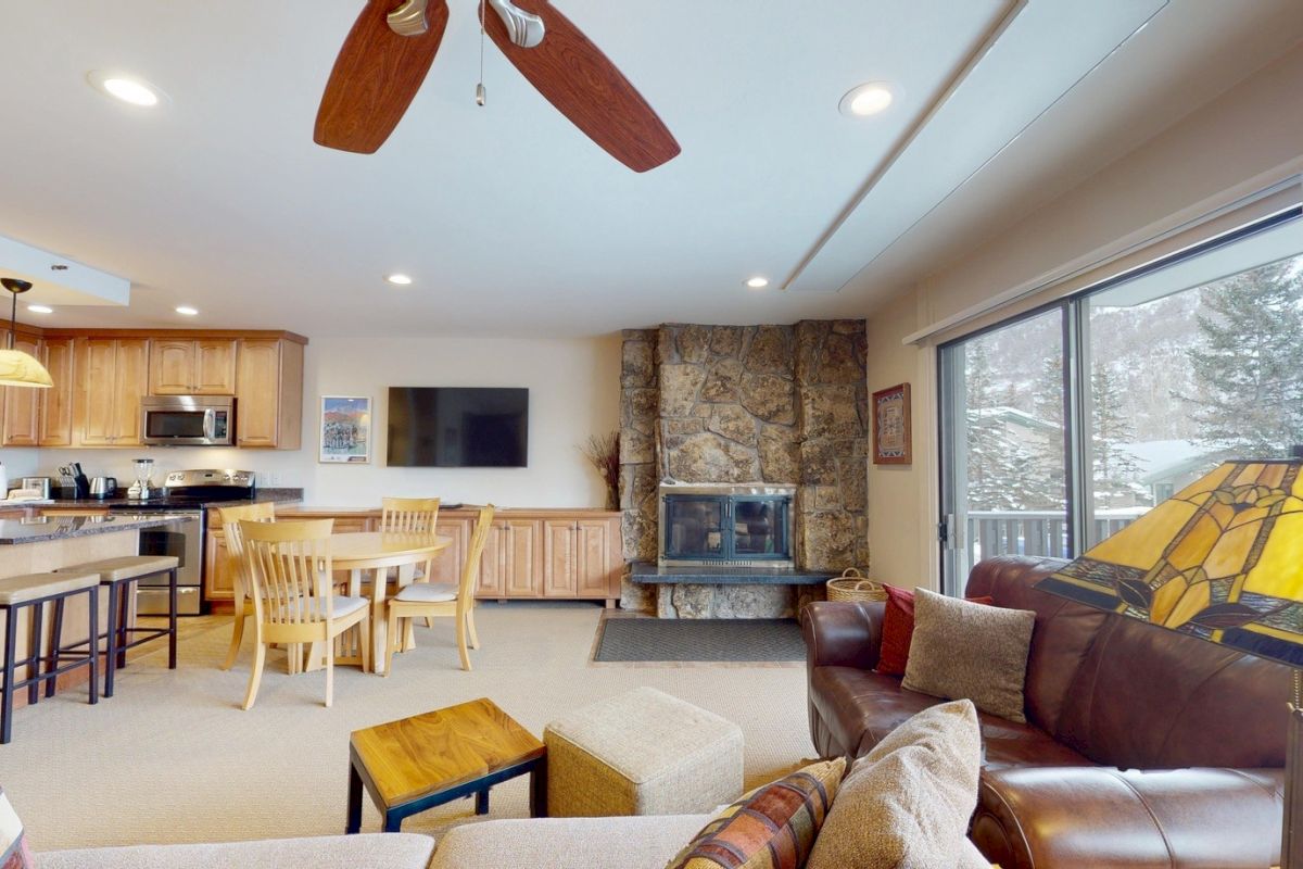 The image shows a cozy living space with a kitchen, dining area, stone fireplace, and large windows, perfect for relaxation.