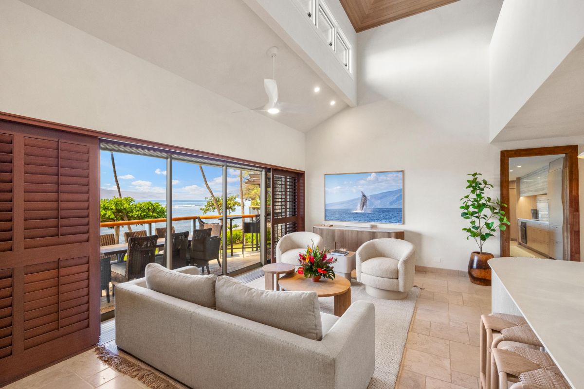 A bright, open living room with beige sofas, a coffee table, tropical outdoor balcony, potted plants, and a doorway leading to a deck with ocean views.