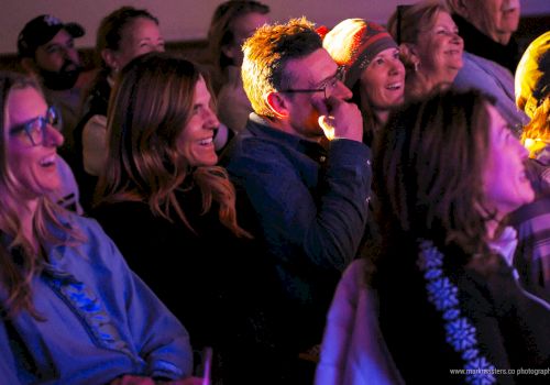 A group of people in a dim, colorful venue watching a performance, smiling and laughing as they sit close together.