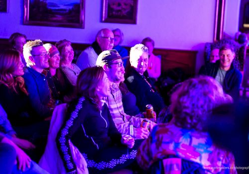 A group of people seated indoors, watching something on stage; the room is bathed in blue-purple lighting.