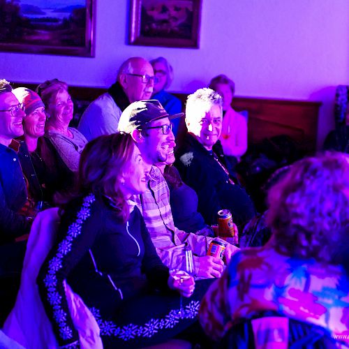 A group of people seated indoors, watching something on stage; the room is bathed in blue-purple lighting.