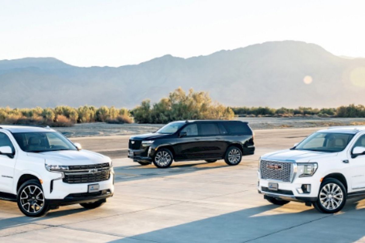 Three large SUVs parked on a sunlit open lot with mountains in the background, white and dark-colored vehicles facing different directions.