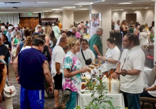 Crowd at a busy indoor market or expo tasting booths, people chatting, sampling foods and drinks, vendors behind tables.