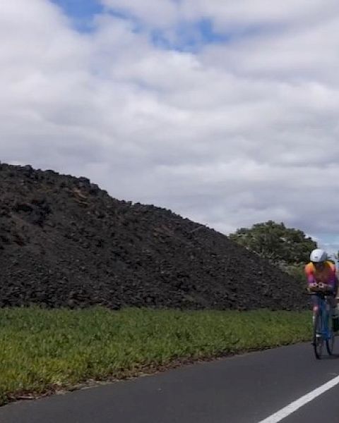 Cyclists ride along a road beside a large dark stone wall and green fields under a cloudy sky.