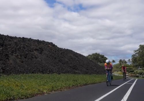 Cyclists ride along a road beside a large dark stone wall and green fields under a cloudy sky.